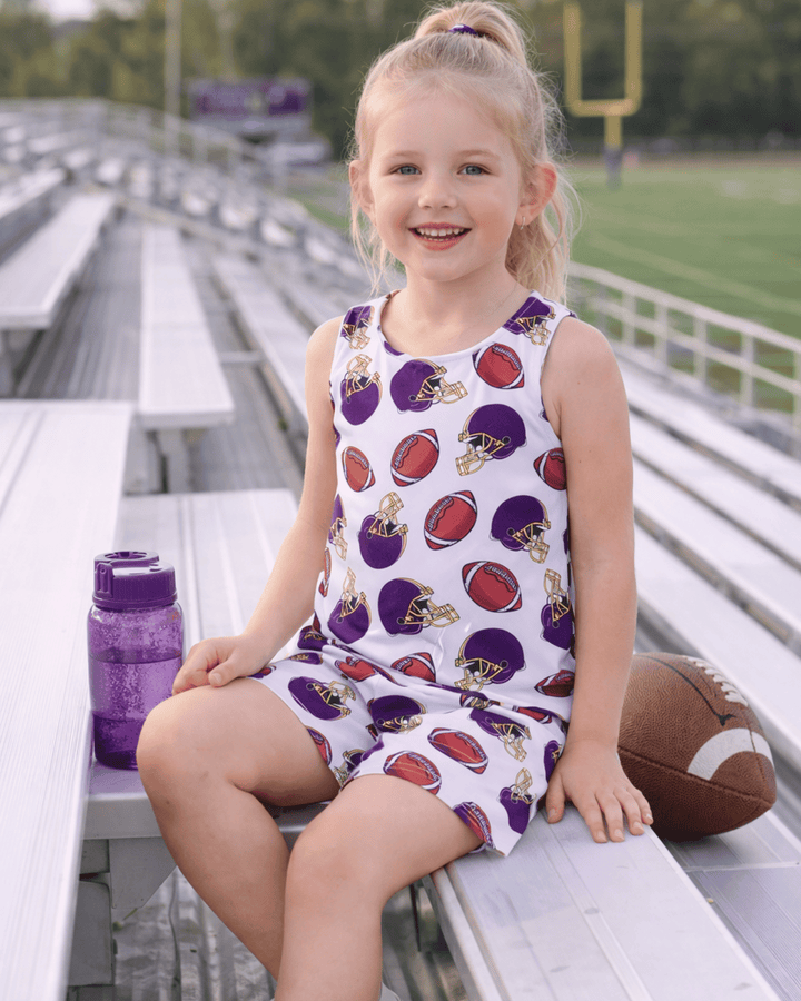 Young girl sitting on bleachers wearing an outfit with sports-themed print, with a football and water bottle nearby.