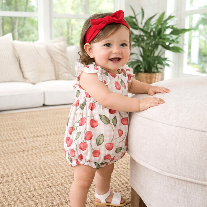 Baby in a cherry-patterned romper standing on a beige rug with a white couch and plant in the background.