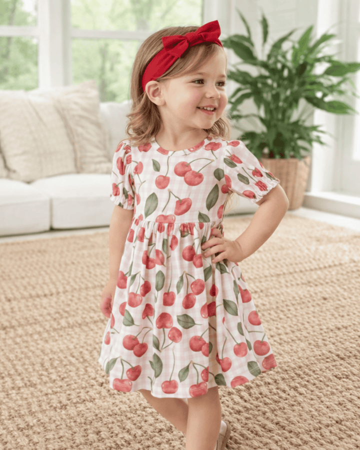 Child wearing a cherry-patterned dress with a red headband in a home setting.