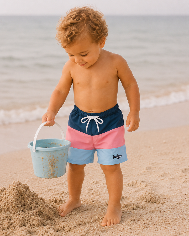 Child in colorful swim shorts playing on a beach with a bucket