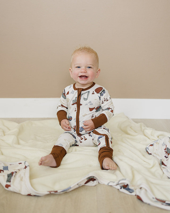 Baby wearing a patterned onesie sitting on a beige blanket against a beige wall.
