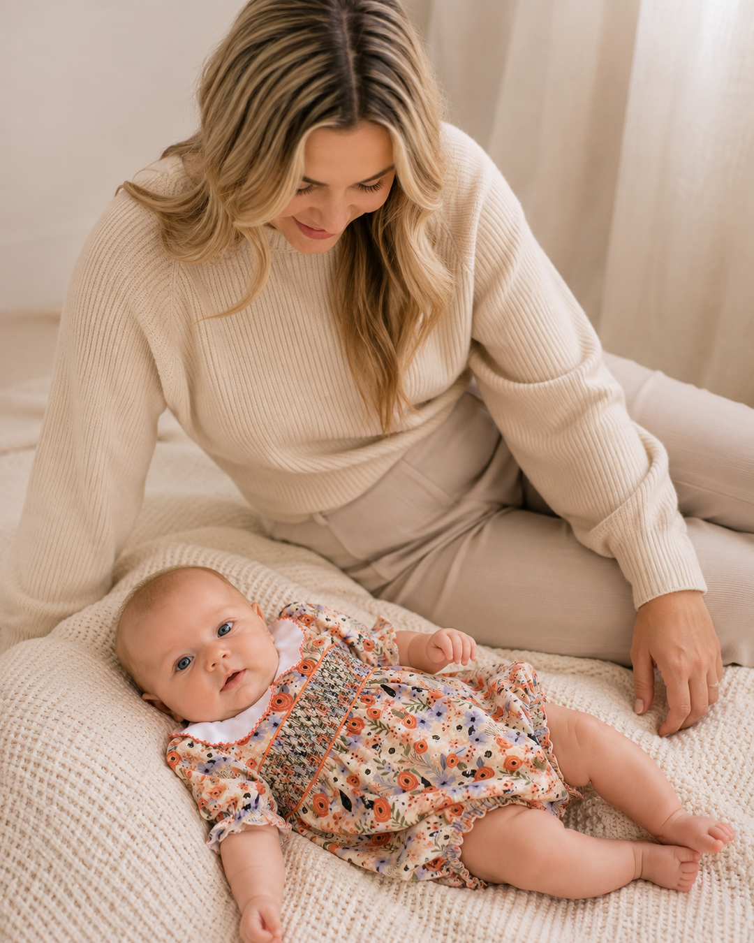 A woman in a cream ribbed sweater and beige pants sits on a textured cream blanket next to a baby wearing a floral long-sleeve romper. The scene is shot in a warm, soft-focus indoor setting.