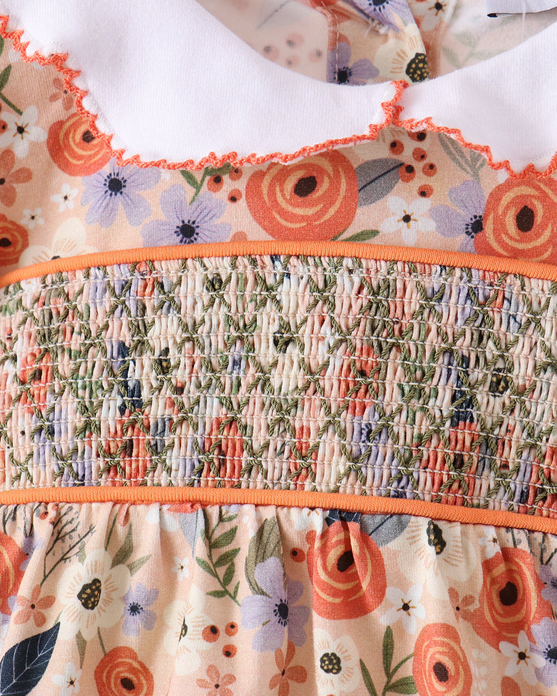 Close-up of a floral dress or top with a smocked waist panel, featuring orange, purple, green, and cream tones, with a white ruffled collar.