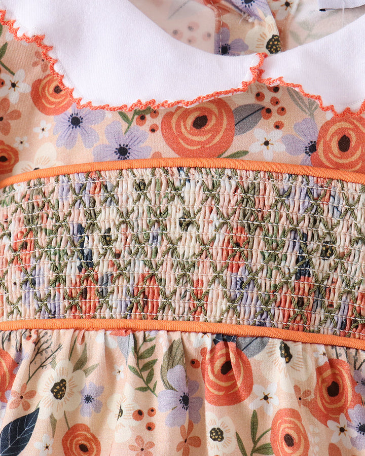 Close-up of a floral dress or top with a smocked waist panel, featuring orange, purple, green, and cream tones, with a white ruffled collar.
