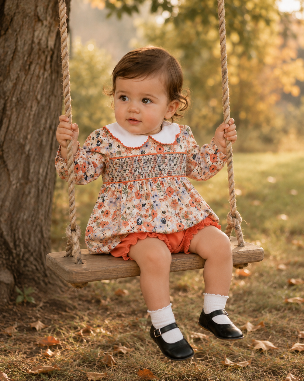 A toddler girl sits on a wooden swing outdoors in a park, wearing a long-sleeve floral smocked dress with a white Peter Pan collar, orange ruffled skirt, white socks, and black Mary Jane shoes.