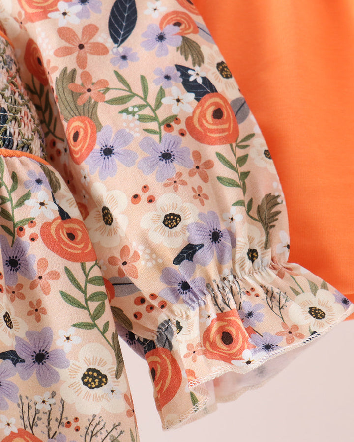 Close-up of a floral-patterned blouse with ruffled sleeves and a solid orange fabric panel, shown against a light pink background.
