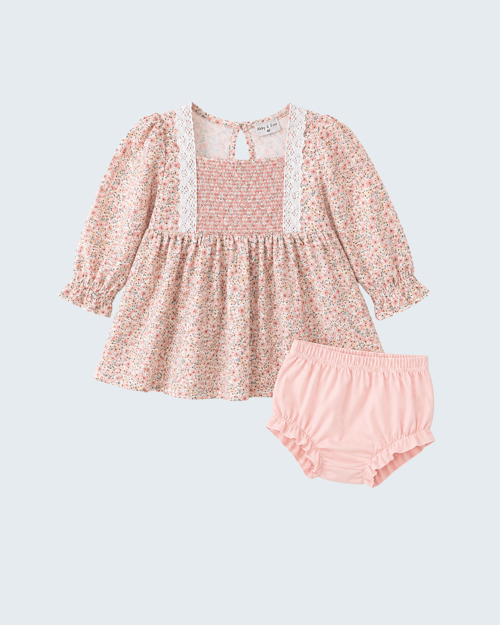 A toddler girl's two-piece outfit set featuring a long-sleeve smocked dress in a pink and white floral print with white lace trim, paired with matching light pink ruffled bloomers, shown on a light gray background.