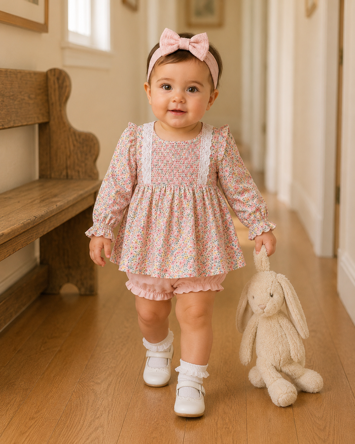 A toddler girl wearing a pink floral long-sleeve dress, matching pink ruffle shorts, a pink bow headband, and white shoes is walking down a wooden hallway while holding a beige plush bunny toy.