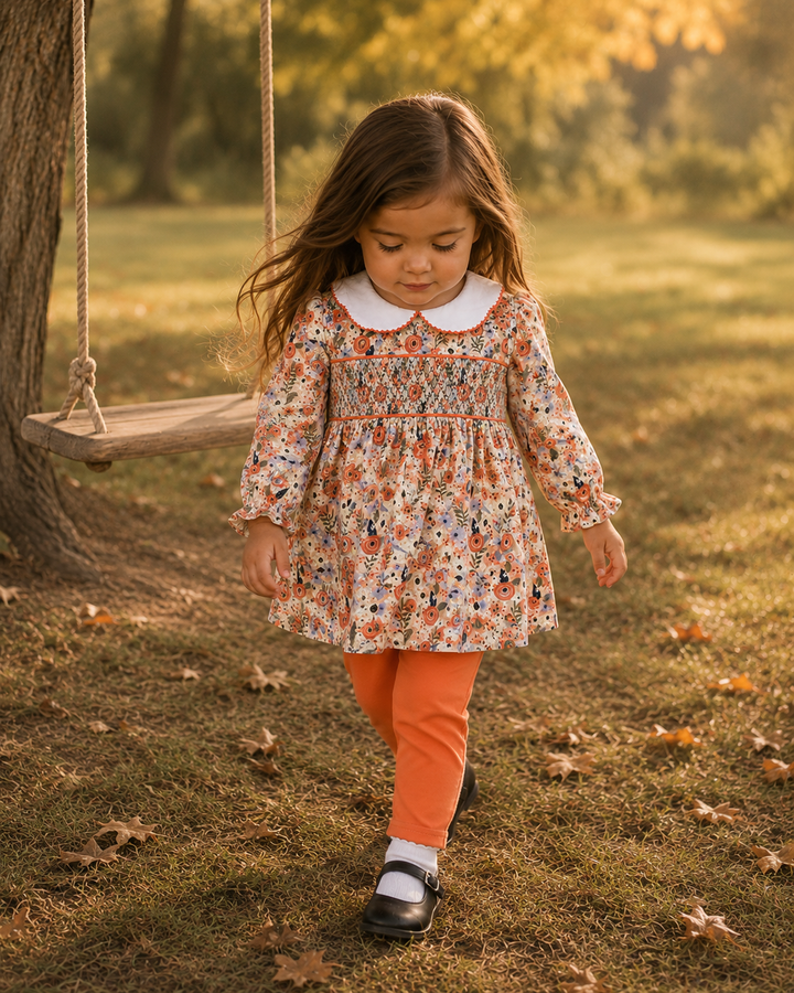 A young girl walking outdoors in a park near a swing, wearing a long-sleeve floral smocked dress with a white Peter Pan collar, bright orange leggings, white socks, and black Mary Jane shoes.