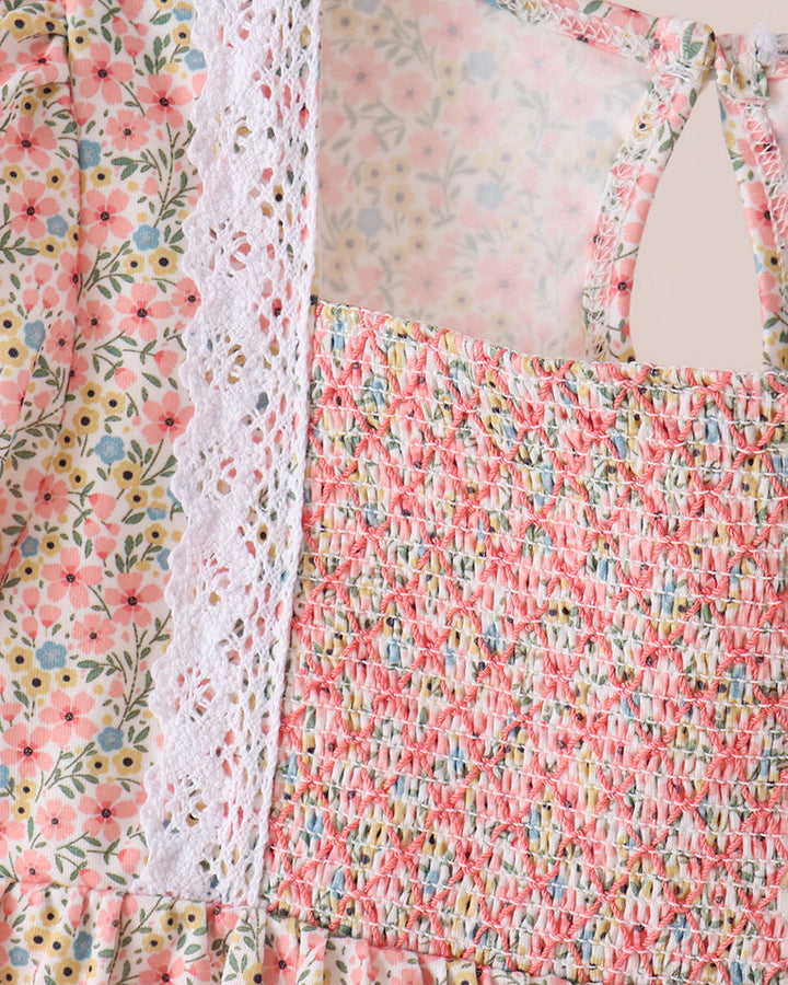 A close-up of a floral smocked dress with a white lace panel and thin shoulder straps, shown against a light beige background.