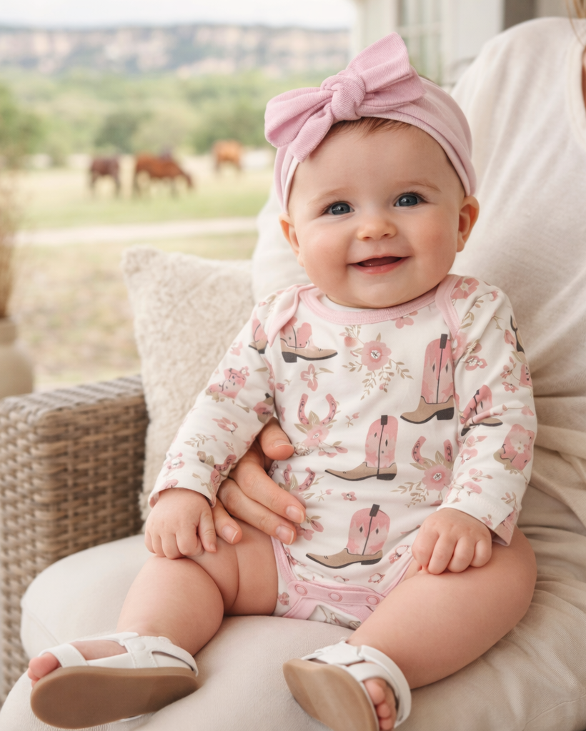 Baby wearing a pink outfit with cowboy boot prints, sitting on a person's lap outdoors.