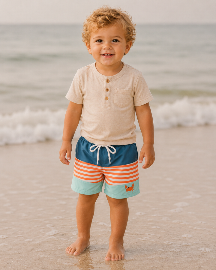 Child wearing a beige shirt and striped shorts standing on a beach with ocean waves in the background