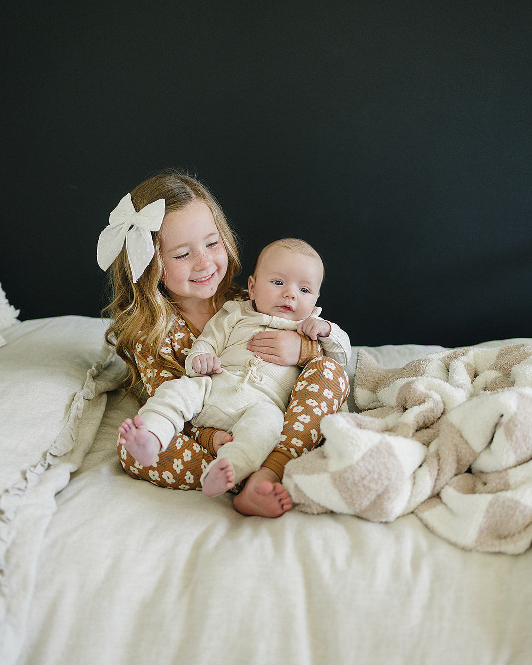 Two children, an older one holding a baby, sitting on a bed with a dark background.