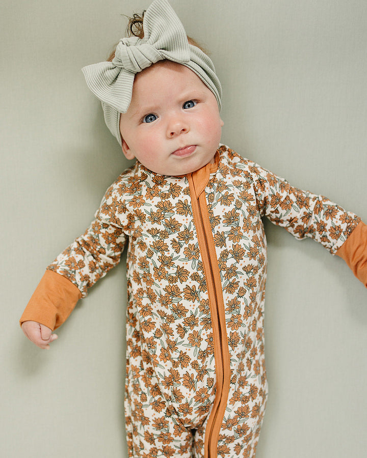 Baby wearing a floral onesie with orange accents and a matching headband against a plain background