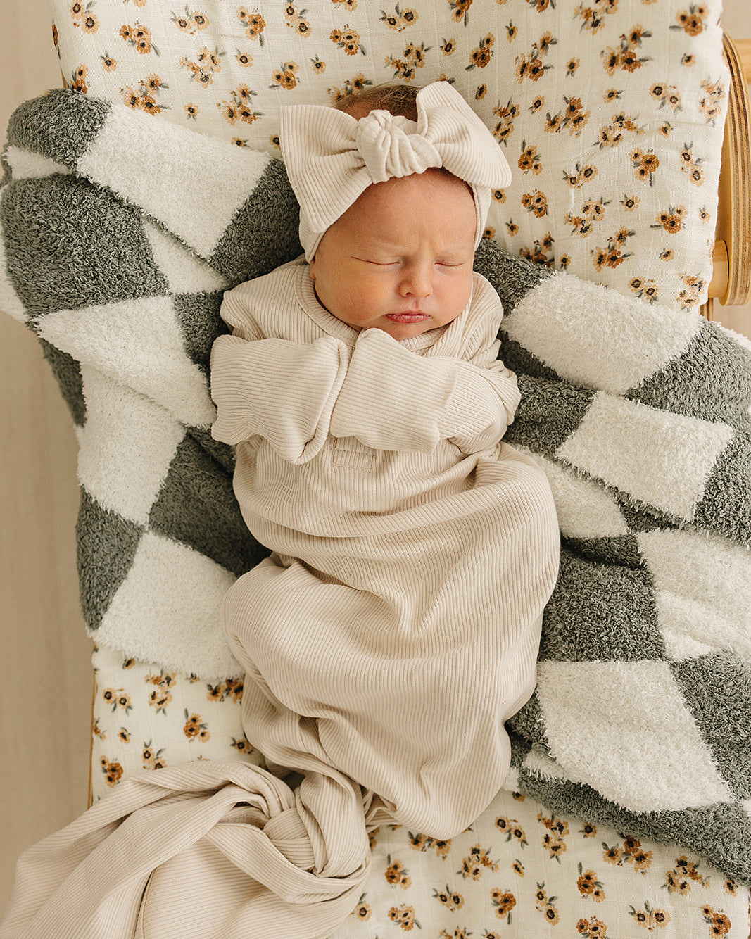 Newborn baby wrapped in a beige blanket with a matching headband, sitting on a floral-patterned chair.
