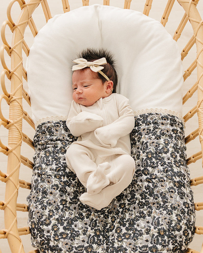 Newborn baby in a wicker crib with a floral blanket and white headband.