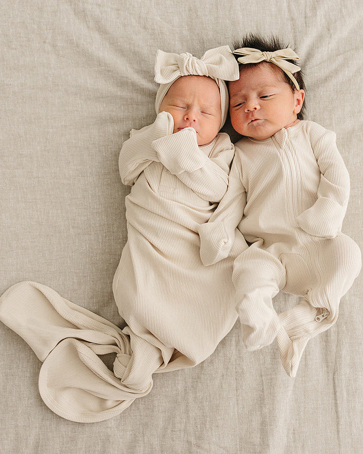 Two newborn babies wrapped in white swaddles with matching headbands on a beige blanket.