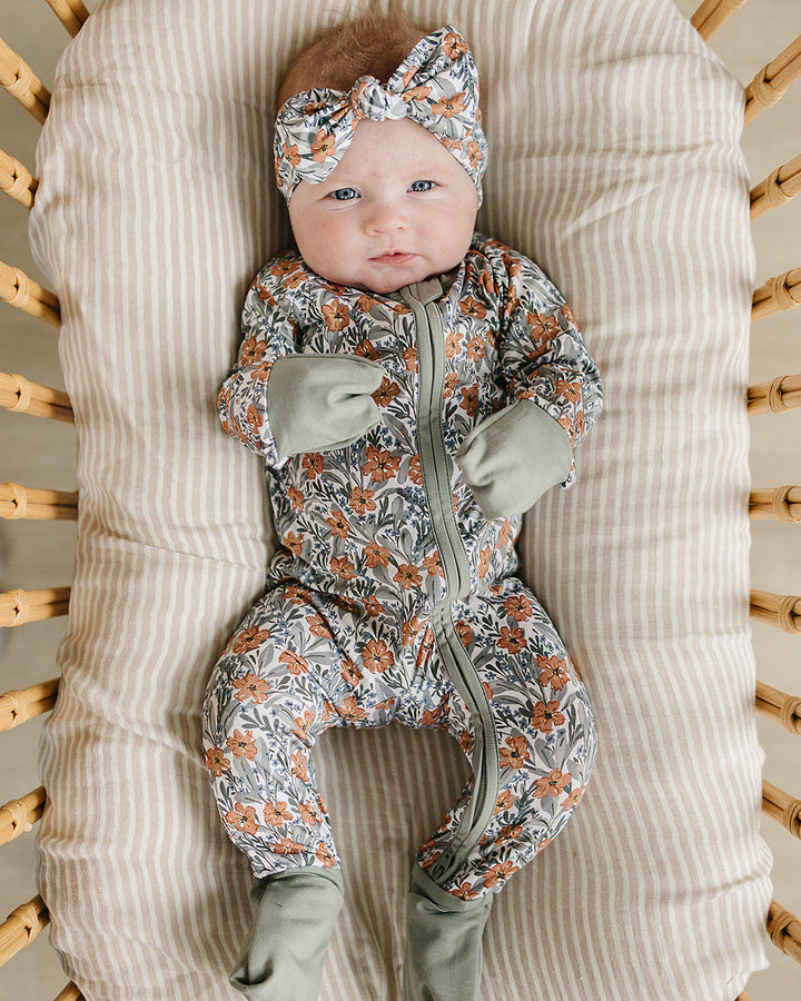 Baby in a floral outfit and green gloves lying on a textured pillow in a crib.