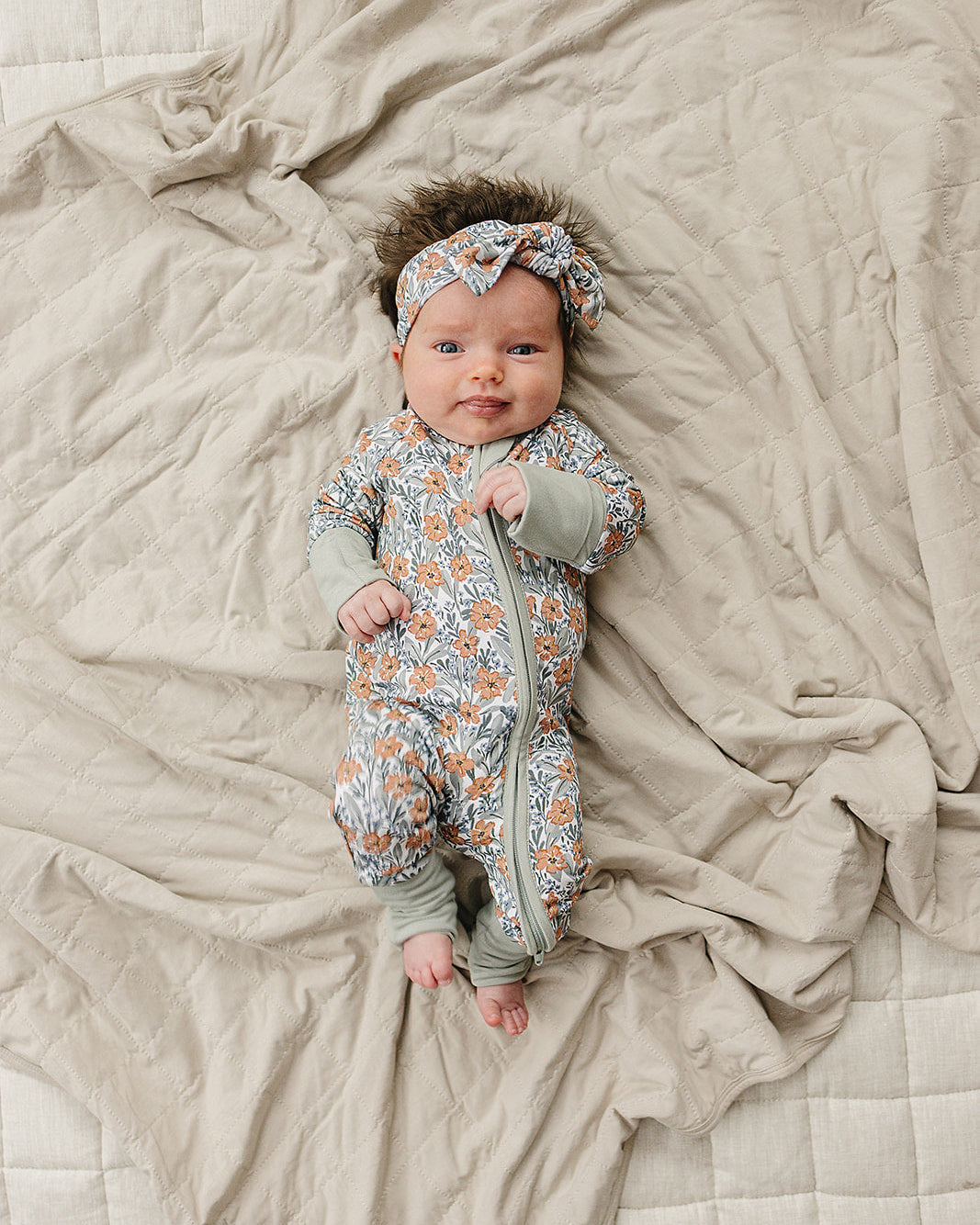 Newborn baby in a floral onesie and headband lying on a soft blanket.