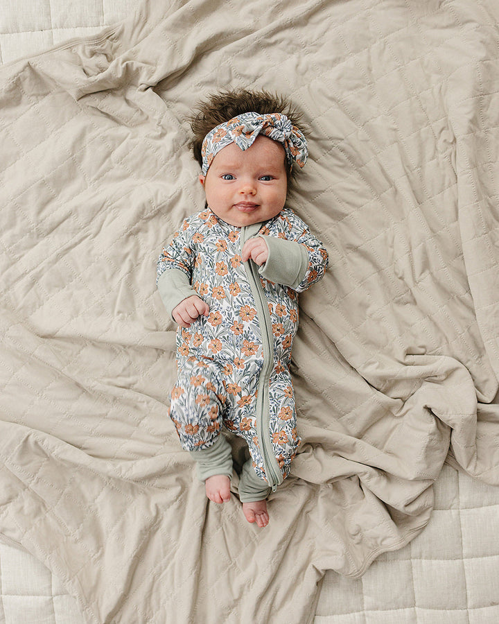 Newborn baby in a floral onesie and headband lying on a soft blanket.
