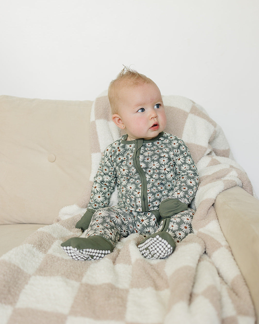 Baby in a floral onesie sitting on a beige couch with a checkered blanket