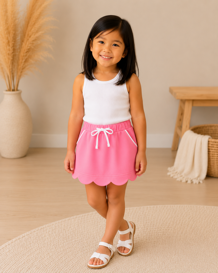 A young girl smiling and posing in a bright indoor room, wearing a white sleeveless tank top and a pink skorts with a scalloped hem and white drawstring.