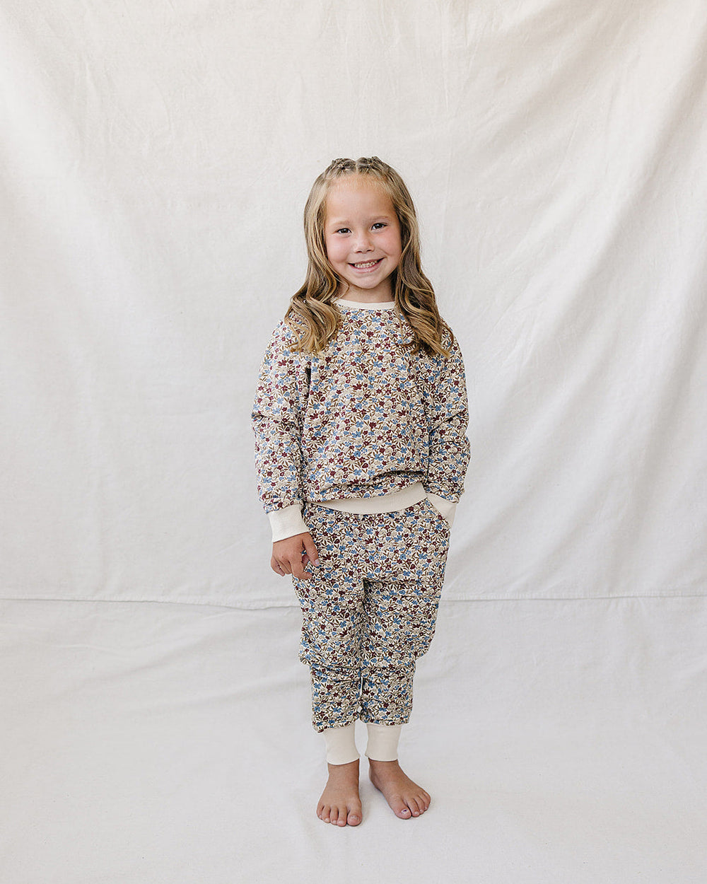 A young girl stands barefoot against a white fabric backdrop, wearing a matching long-sleeve sweatshirt and jogger pants set with a dense small floral print on a light base.