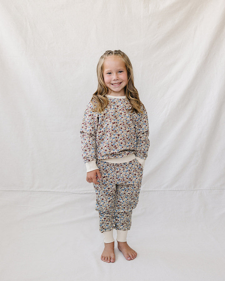 A young girl stands barefoot against a white fabric backdrop, wearing a matching long-sleeve sweatshirt and jogger pants set with a dense small floral print on a light base.