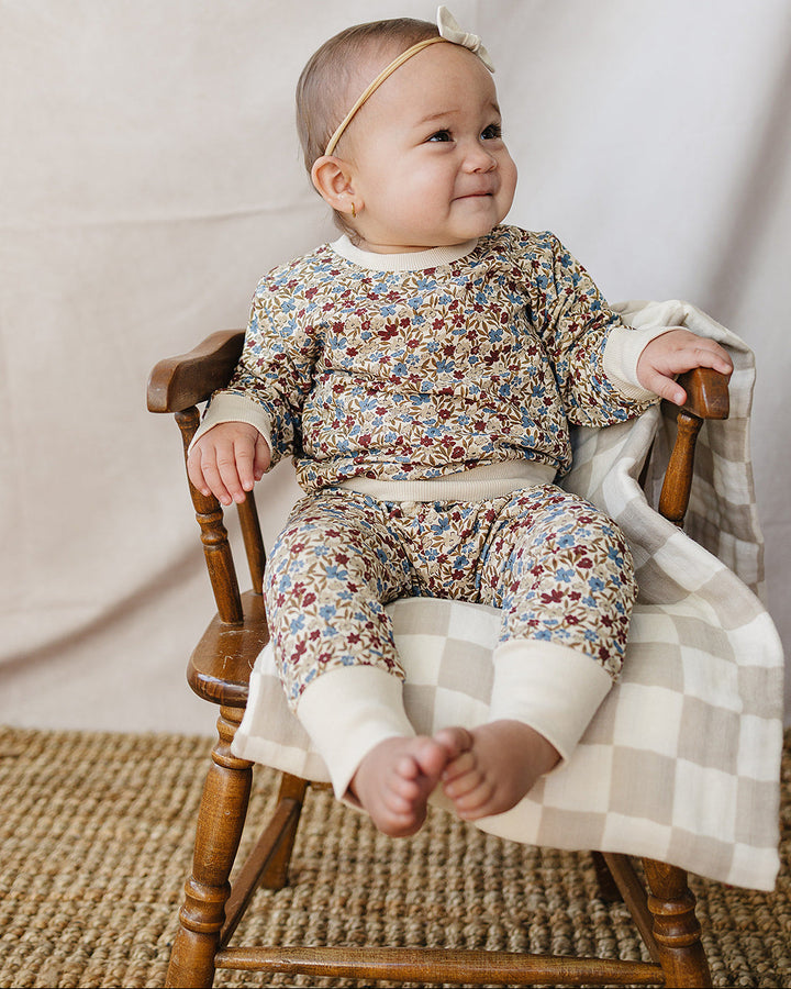 A baby wearing a matching floral print romper and pants set with cream cuffs, sitting on a wooden chair with a checkered blanket against a draped cream fabric backdrop.