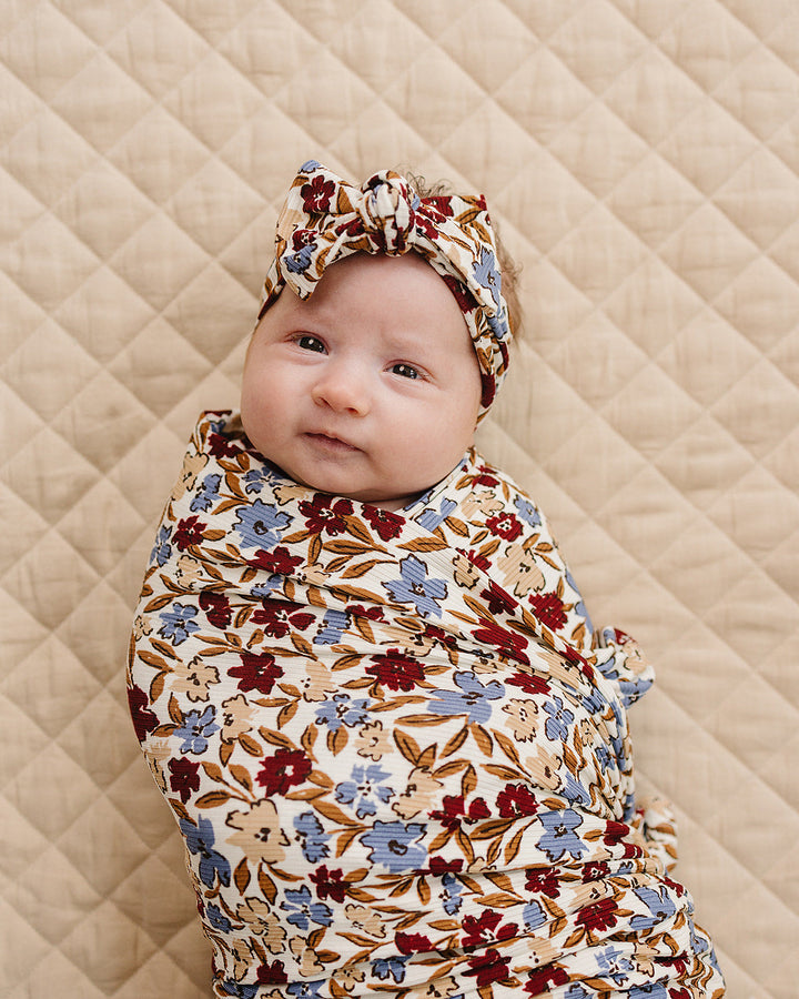 Baby wrapped in a floral swaddle and headband on a quilted beige surface