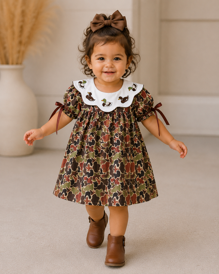 Child wearing a floral dress with a white collar and brown shoes, standing indoors.