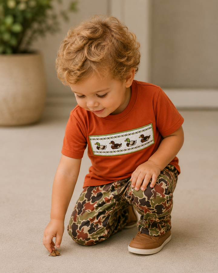 Child wearing a red shirt with a graphic design and patterned pants, sitting on the floor.