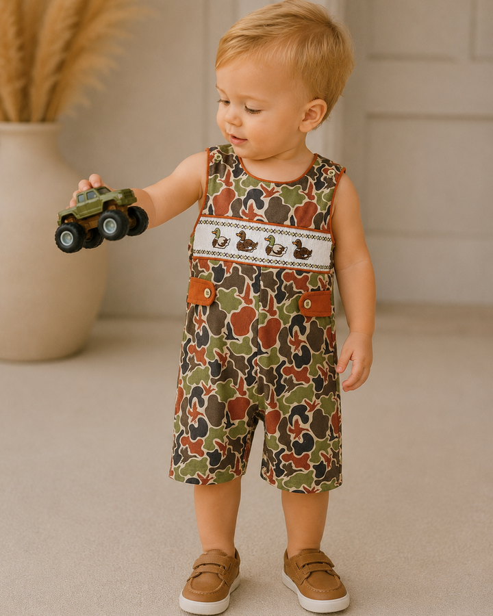 Child wearing a camouflage romper holding a toy car indoors.