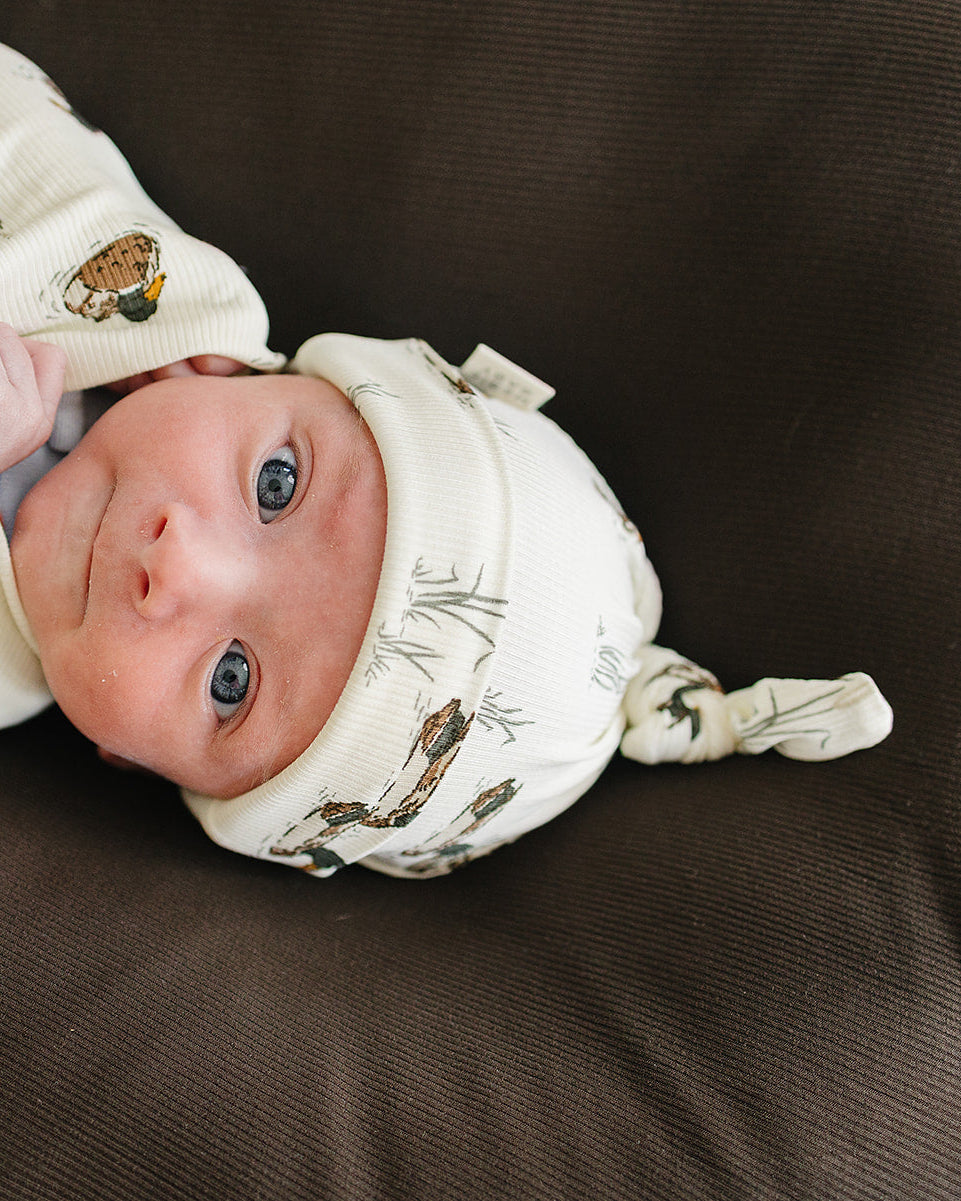 Newborn duck-print knot hat in white ribbed knit, baby lying down