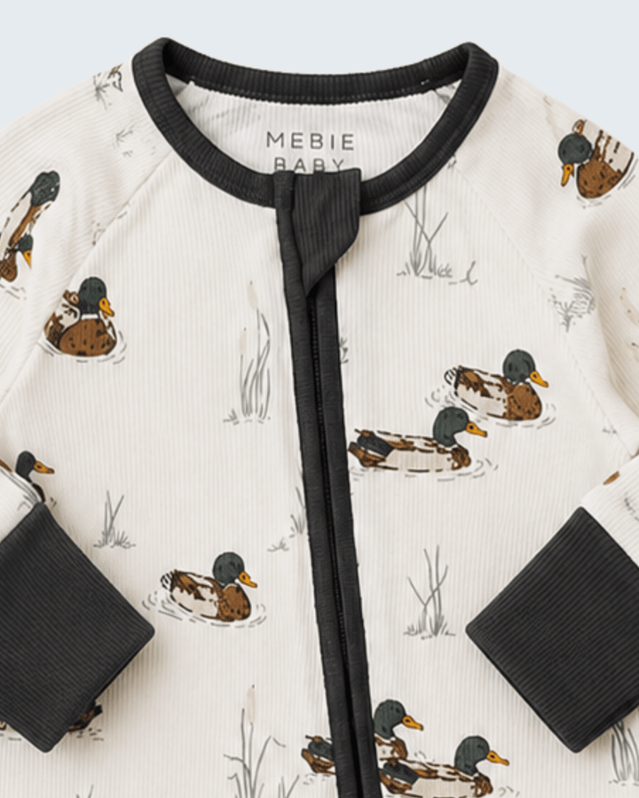 Close-up of a baby zip-up romper by Mebie Baby, featuring an off-white ribbed fabric with a printed pattern of mallards and reeds, black ribbed collar, cuffs, and front zipper.