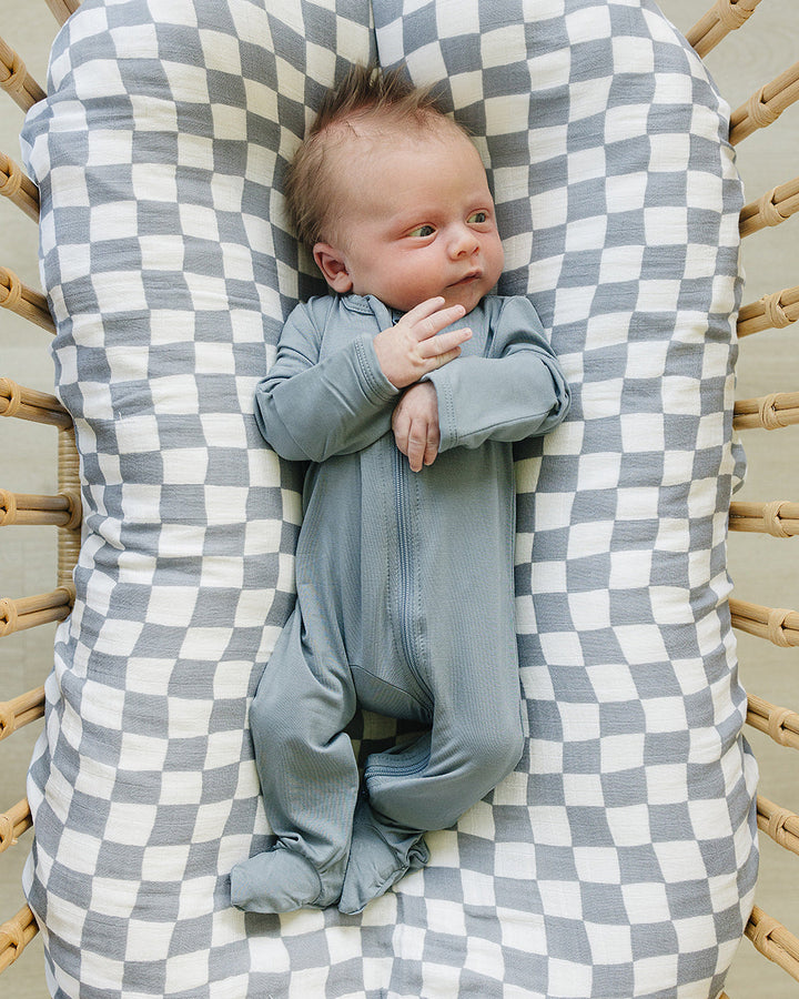 Newborn baby in a gray outfit lying on a checkered cushion in a wooden crib