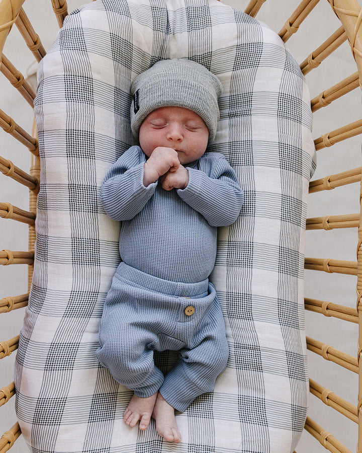 A sleeping newborn baby wearing a light blue ribbed knit bodysuit and matching beanie hat, lying on a gray and white plaid bassinet cushion inside a wicker basket.