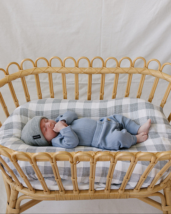 A newborn baby wearing a light blue knit onesie and matching beanie hat, sleeping peacefully inside a natural rattan bassinet with a blue and white plaid cushion.