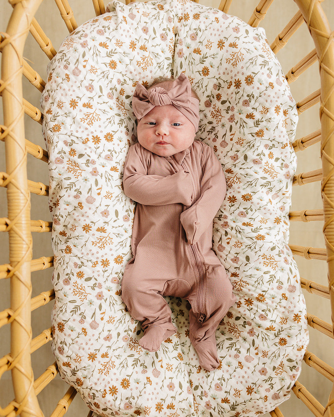 Newborn baby in pink outfit and headband lying on a floral-patterned cushion inside a wicker basket.