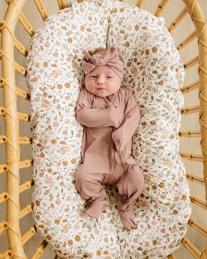 Newborn baby in pink outfit and headband lying on a floral-patterned cushion inside a wicker basket.