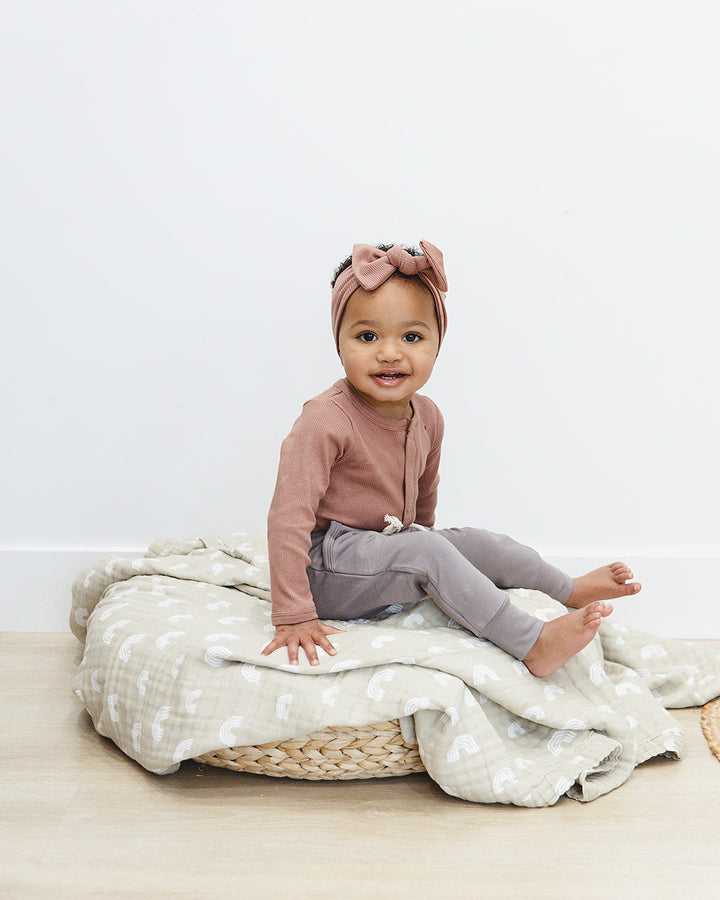Baby sitting on a woven mat with a white blanket, wearing a pink top and gray pants.
