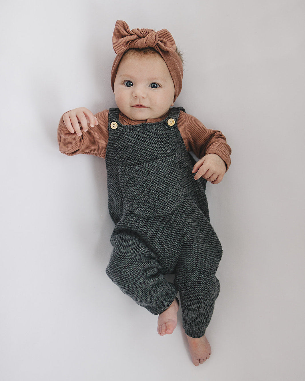 Baby wearing a brown top and gray overalls with a bow headband on a white background