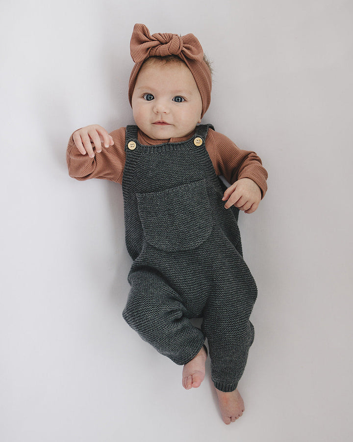 Baby wearing a brown top and gray overalls with a bow headband on a white background