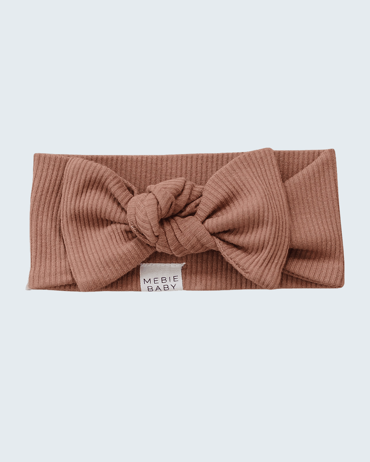A ribbed knit headband in a dusty rose-brown color, featuring a large central knot bow and a small white fabric label that reads &