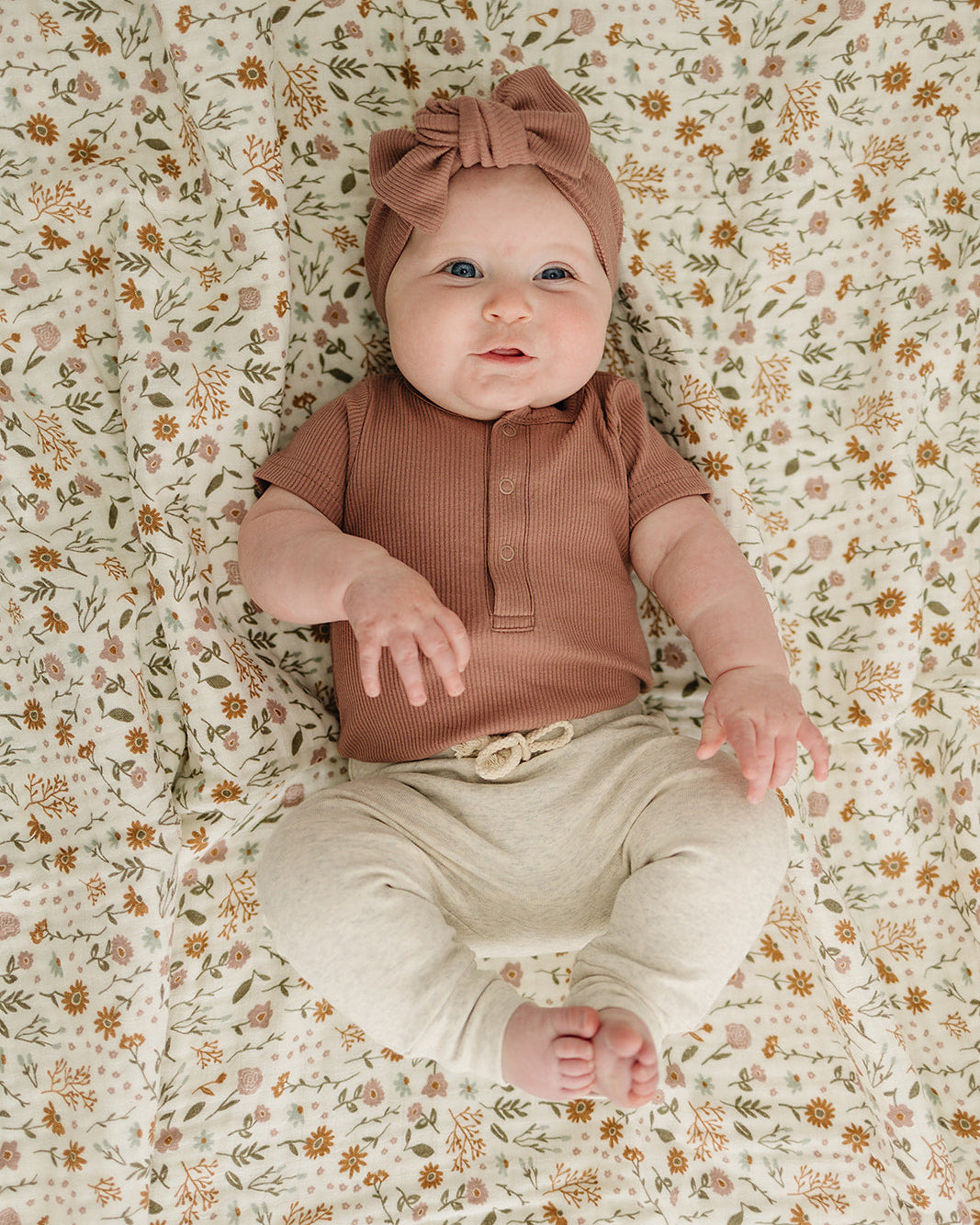 Baby lying on a floral-patterned blanket wearing a brown top and beige pants.