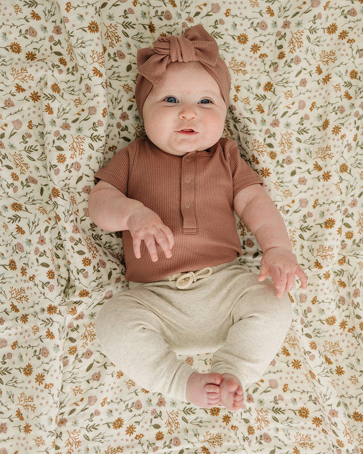 Baby lying on a floral-patterned blanket wearing a brown top and beige pants.