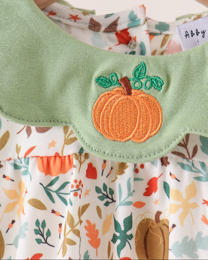 A close-up of a baby dress hanging on a wooden hanger, featuring a light green collar with an embroidered orange pumpkin applique and a white floral print bodice with autumn leaves and pumpkins.