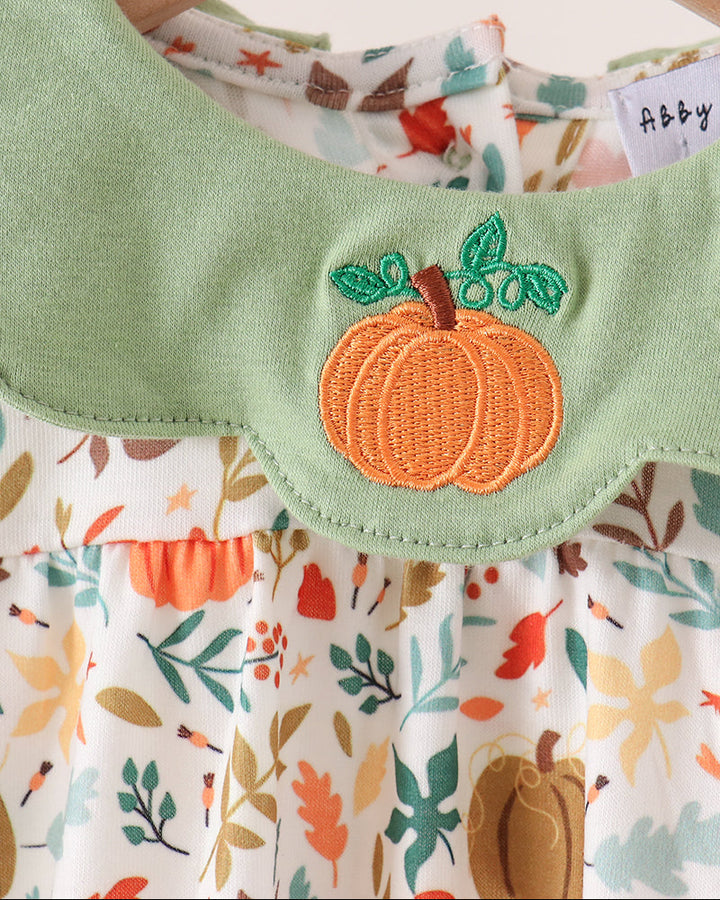 A close-up of a baby dress hanging on a wooden hanger, featuring a light green collar with an embroidered orange pumpkin applique and a white floral print bodice with autumn leaves and pumpkins.