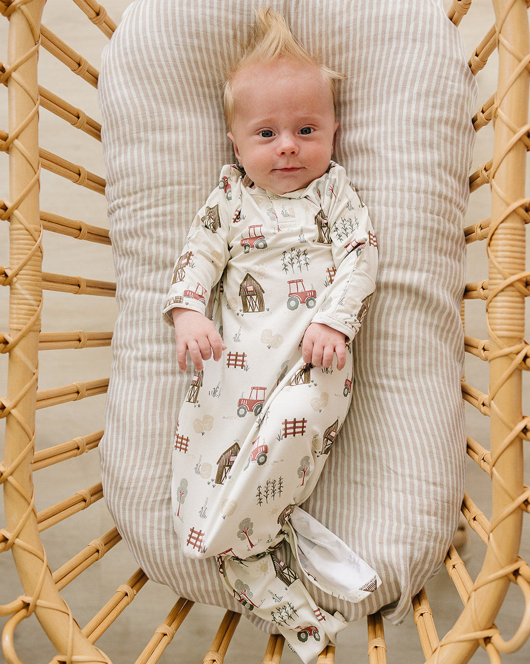 A baby wearing a cream footed sleeper with a farm-themed print of tractors, barns, and fences, lying on a striped cushion inside a wicker chair.