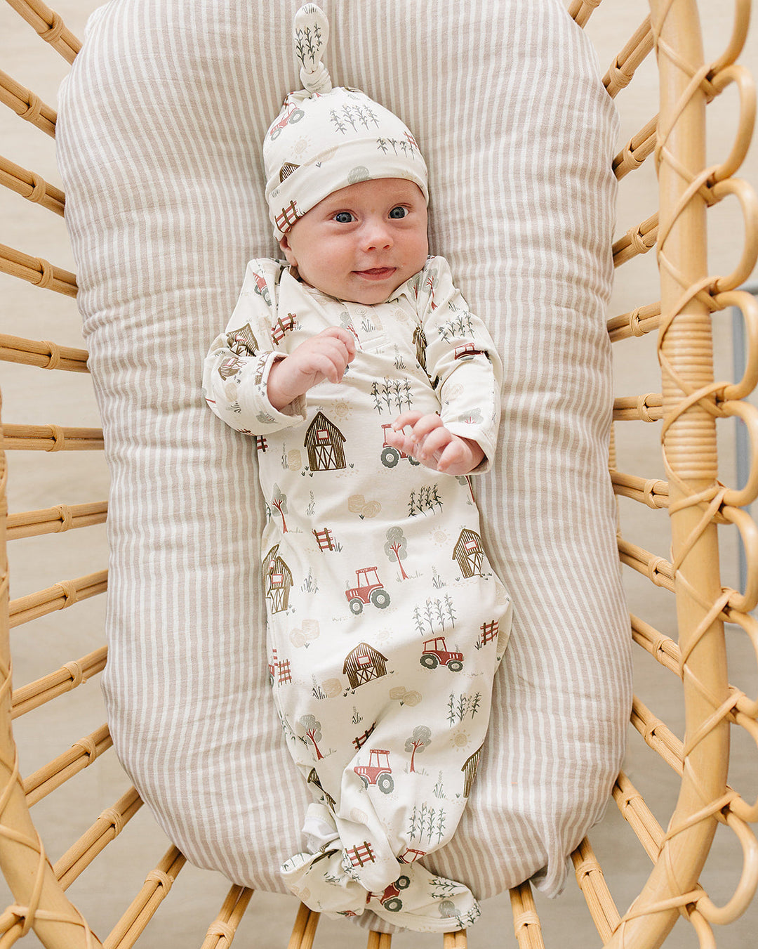 A baby wearing a cream-colored footed sleeper and matching hat with a tractor and barn pattern, lying on a striped cushion inside a wicker chair.